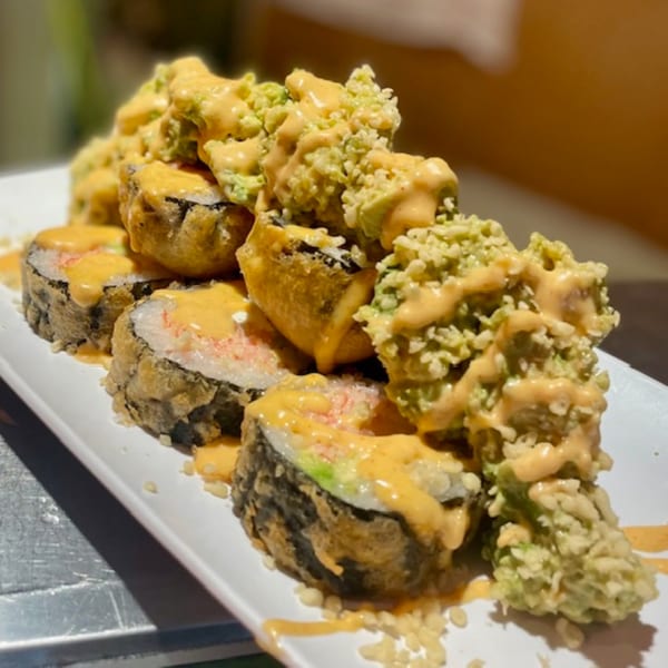 Deep-fried sushi rolls topped with creamy orange sauce and green tempura flakes on a white plate.