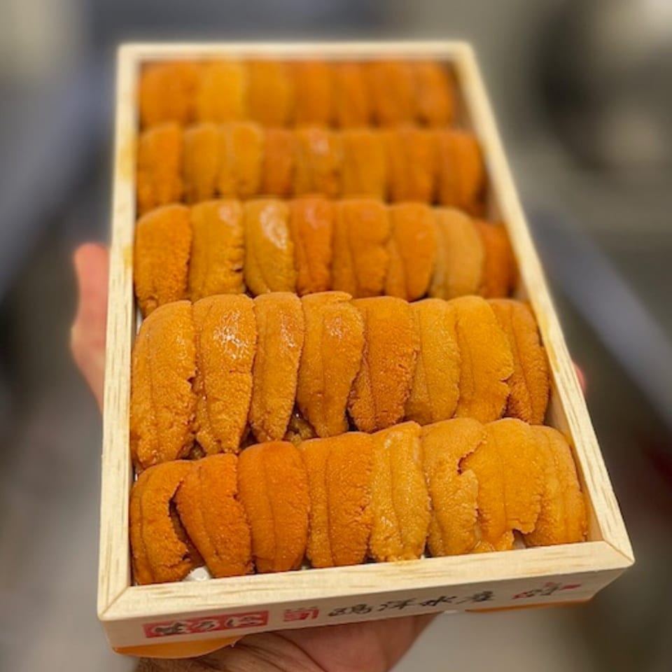 A hand holds a wooden box filled with rows of bright orange uni (sea urchin roe).