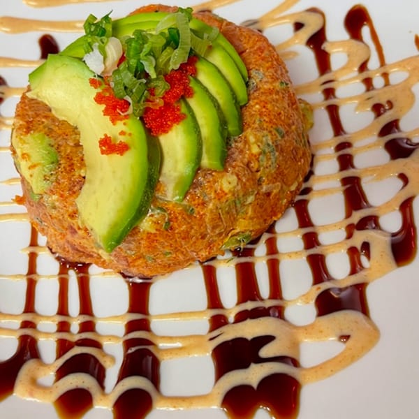 Sushi rice cake topped with sliced avocado, red tobiko, and green onions, drizzled with sauces.